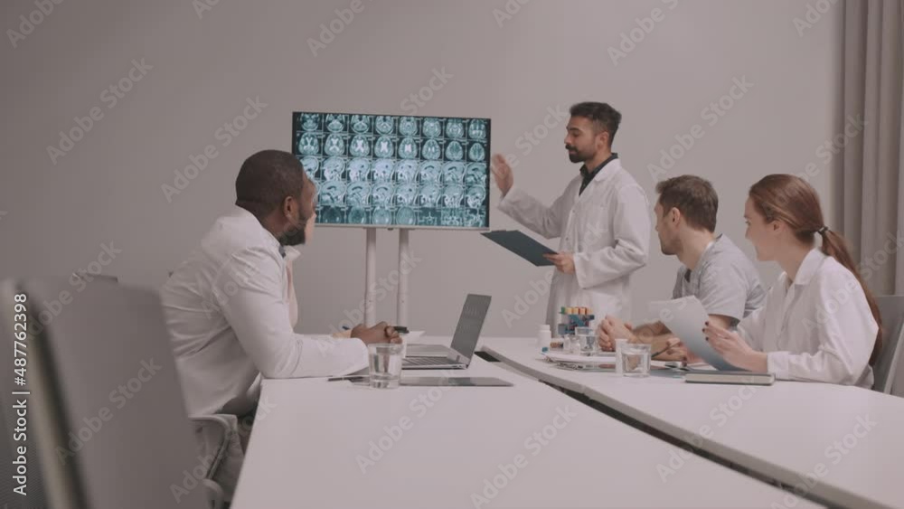 Medium shot of group of multiethnic male and female neurologists having meeting in conference room. Biracial man talking about brain x-ray of patient in front of colleagues Medium shot of group of multiethnic male and female neurologists having meeting in conference room. Biracial man talking about brain x-ray of patient in front of colleagues