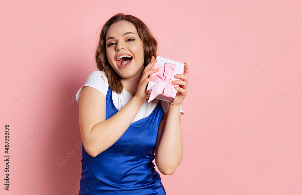 Excited young beautiful girl with long curly hair holding small gift box isolated on pink background. Concept of emotions, beauty, fashion, holidays Excited young beautiful girl with long curly hair holding small gift box isolated on pink background. Concept of emotions, beauty, fashion, holidays