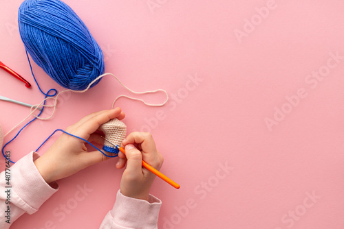 Children's hands in the process of crocheting toys from blue and beige yarn. Pink background. Needlework, hobbies, craft training. The development of fine motor skills. Flat lay, copy space. photo