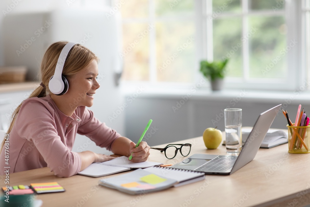 Laughing cute blonde european young girl in headphones study at home look at pc in kitchen interior Laughing cute blonde european young girl in headphones study at home look at pc in kitchen interior