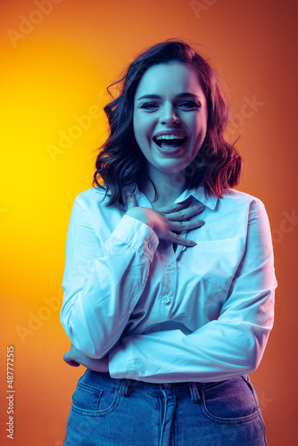 Portrait of young beautiful girl with long curly hair laughing isolated on orange background in neon light, filter. Concept of emotions, beauty, fashion
