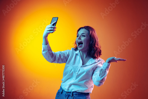 Excited young beautiful girl with long curly hair using smartphone isolated on orange background in neon light, filter. Concept of emotions, beauty, fashion