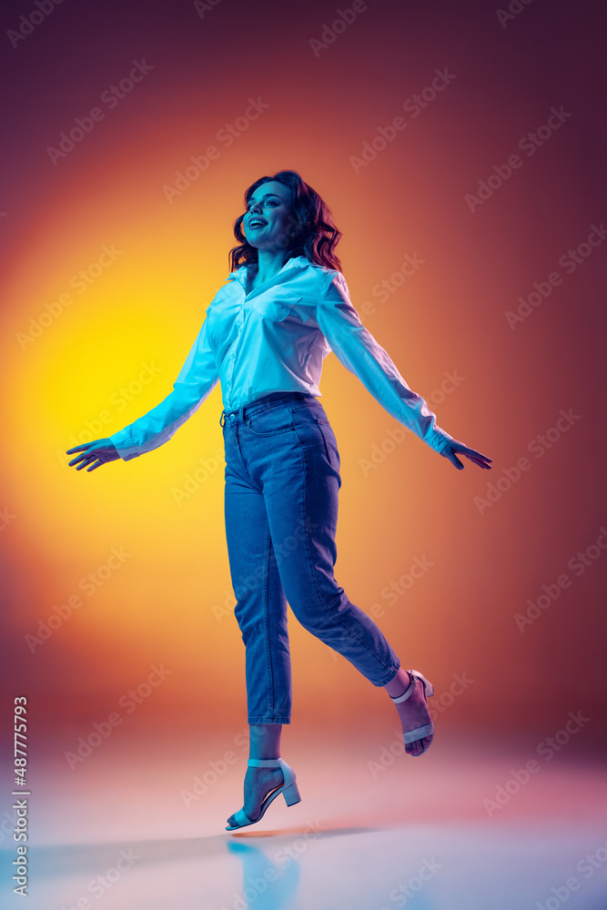 Full-length portrait of happy young beautiful girl with long curly hair jumping isolated on orange background in neon light, filter. Concept of emotions, beauty, fashion