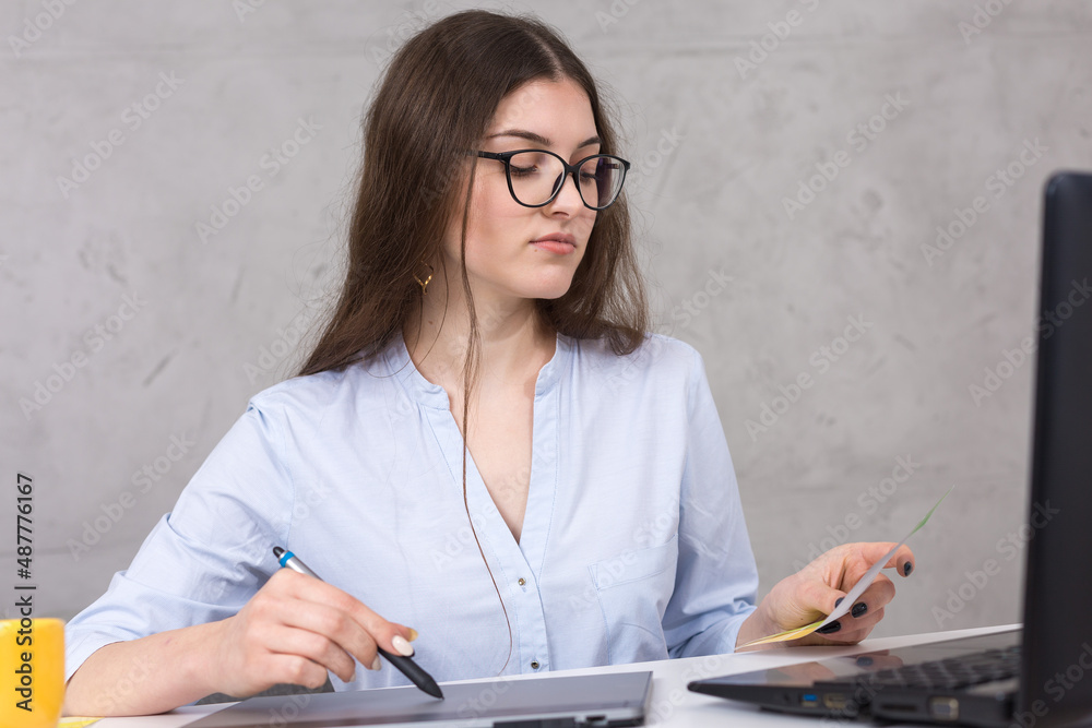 A young designer girl sitting at a table draws on a tablet. Makes business calculations on the computer. A young designer girl sitting at a table draws on a tablet. Makes business calculations on the computer.