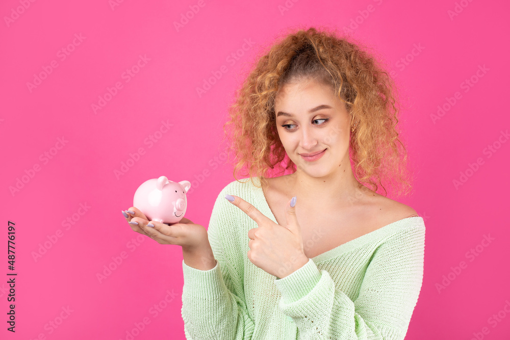 A cute young girl with curly red hair holds a piggy bank, a pink piglet in her hands. The concept of wealth and safety of money. A cute young girl with curly red hair holds a piggy bank, a pink piglet in her hands. The concept of wealth and safety of money.