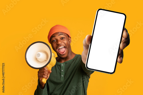 Excited black guy shouting into megaphone, showing big smartphone with empty screen, offering mockup space for ad photo