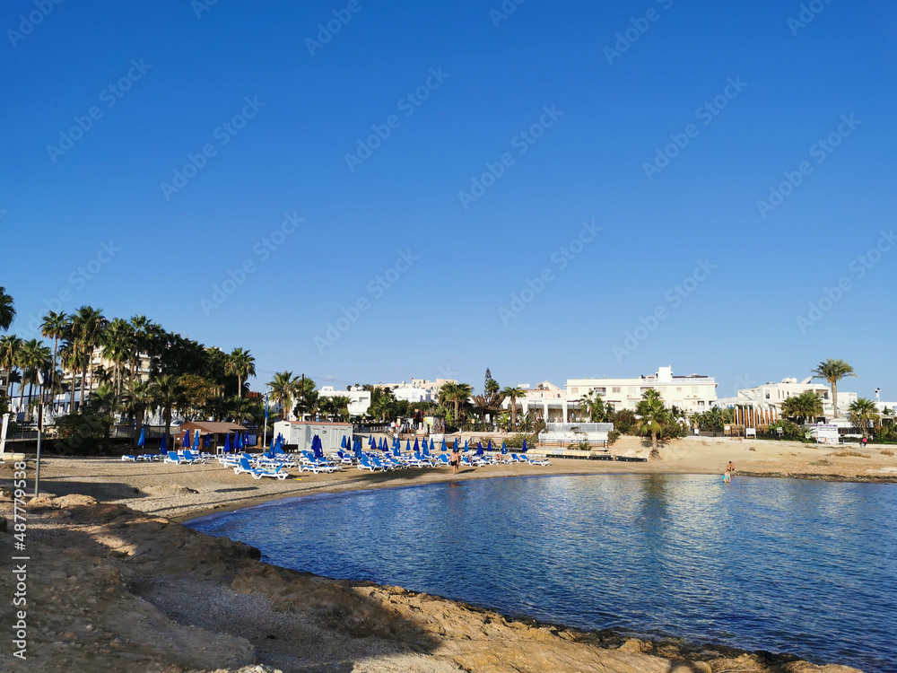 A small sparsely populated sandy beach with sun loungers and umbrellas in the bay of the Mediterranean Sea on a cloudless day.