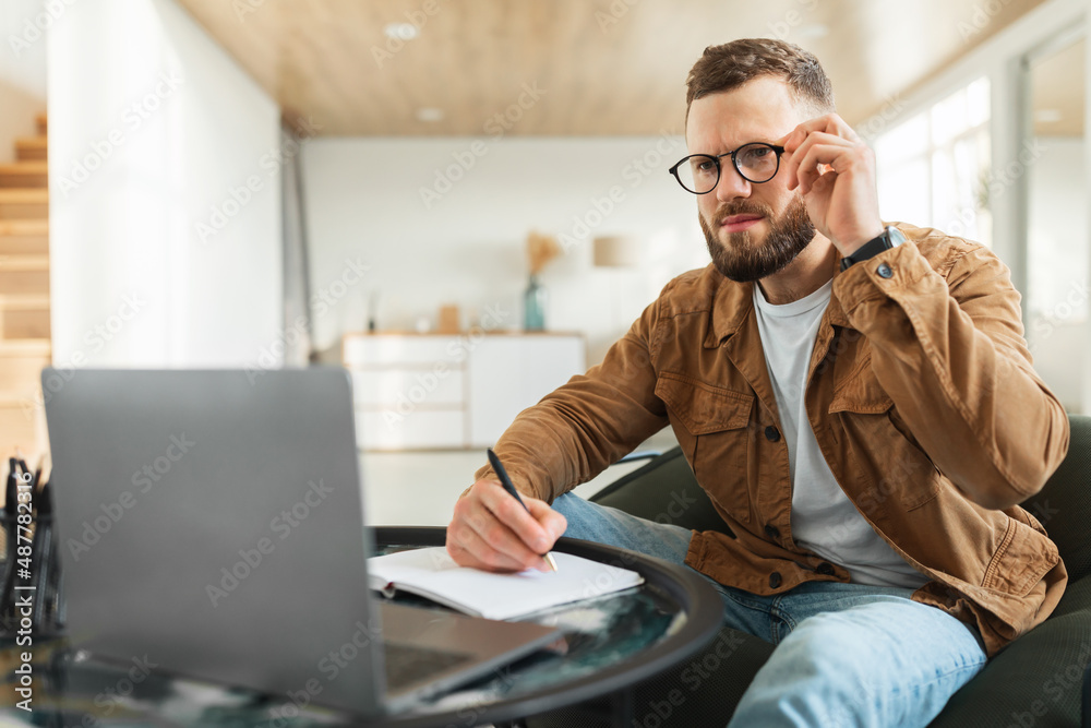 Businessman Looking At Laptop Touching Eyeglasses Taking Notes At Home