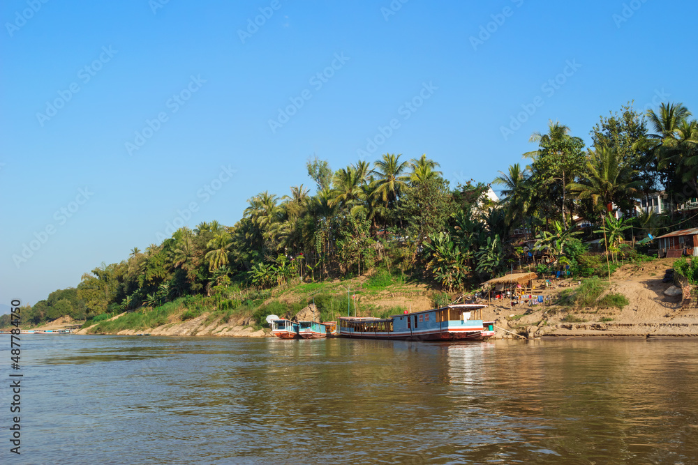 View on riverside with long tail boats, palms trees and houses local people. Mekong River, Luang Prabang, Laos.