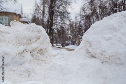 A wide large snowdrift by the road against the backdrop of a city street. On the road lies dirty snow in high heaps. Urban winter landscape. Cloudy winter day, soft light. © Andrei