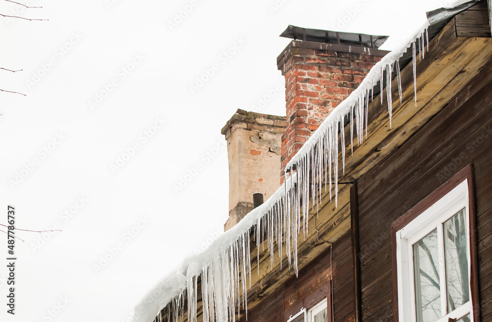 Smooth, transparent icicles hang from the edge of the roof. Against the background of the wooden wall of the old house. Large cascades, even beautiful rows. Cloudy winter day, soft light.