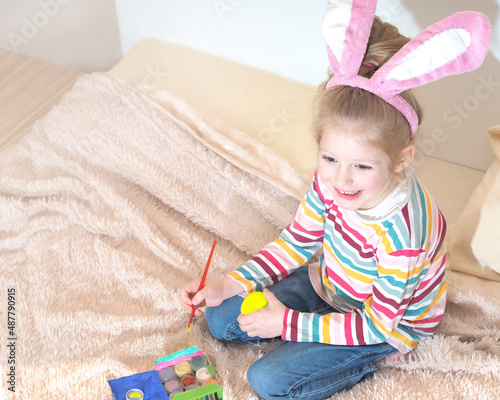 A happy little blonde girl with bunny ears laughs and paints an Easter egg with a paintbrush. Easter preparation. Making Easter decor.