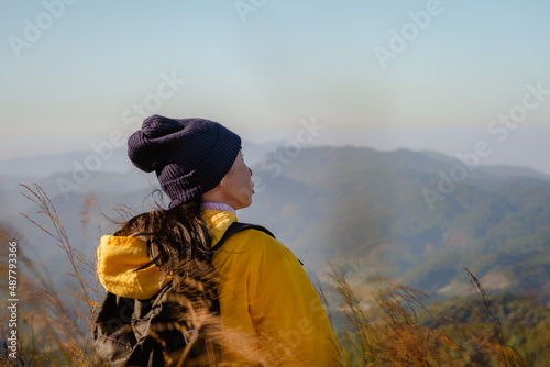 Rear view of a woman in a yellow jacket standing on top of a mountain at sunrise and looking up while enjoying a quiet day in nature breathing in the fresh air. © Pornpimon