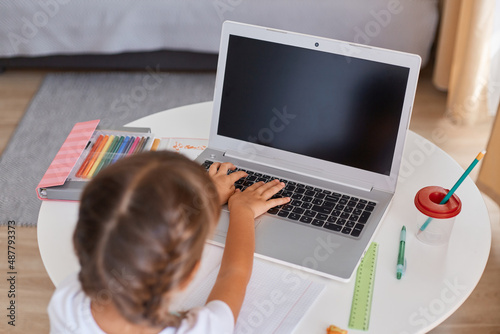 Indoor shot of dark haired female child with braids typing on keyboard, doing her school tasks, distance education, lessons at home, little girl sitting at table and studying on laptop.