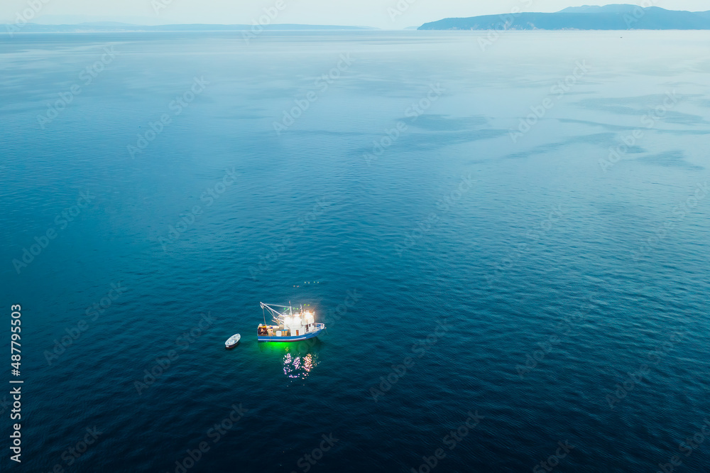 Evening fishing in Adriatic sea with boat drifting on calm water surface.Twilight over sea and distant peninsula with high mountain. Aerial panorama