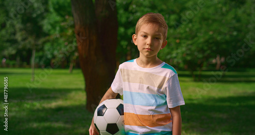 Boy with soccer ball posing camera on nature. Serious little athlete closeup.