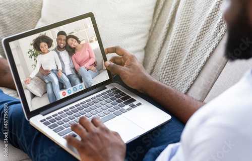 Young black guy communicating to his family online, sitting on sofa with laptop, speaking to friends on webcam