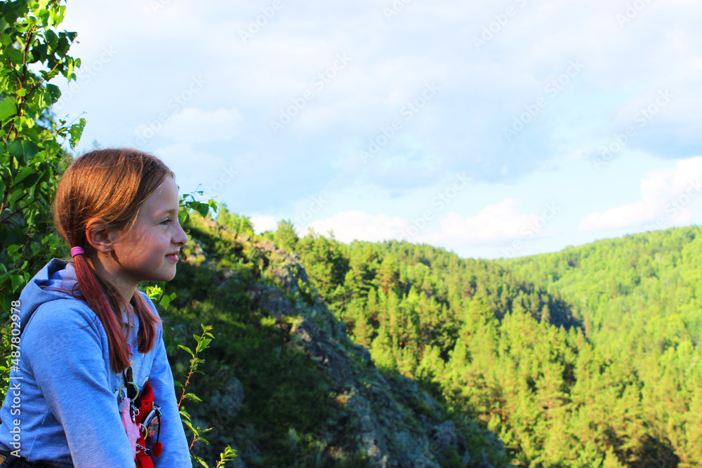 the girl sits alone on the edge of the cliff and looks at the river. the mountains. Forest. Mountain landscape through the foliage of trees. Breathtaking panoramic view from the top of the peak