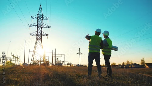 Teamwork industry a concept. Two engineers electric experts in protective helmets discussing the construction of a power line. Silhouette Of Engineers Standing On Field With Electricity Towers.