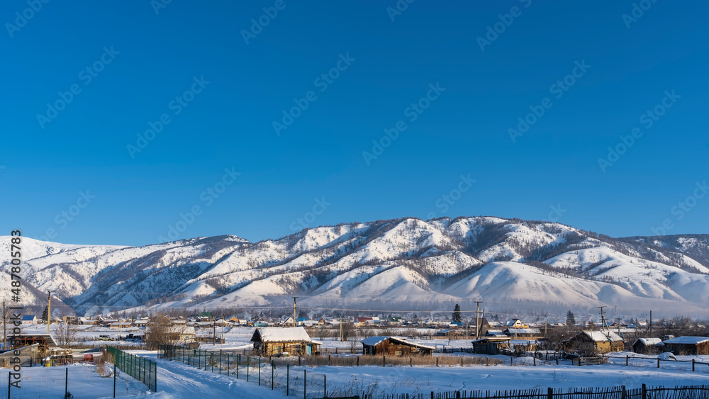 Siberian village in winter. One-story wooden houses, fences, roads are visible. A picturesque snow-covered mountain range against a blue sky. Altai Republic