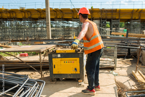 Builder in a hard hat and vest bending rebar on a bending machine at a construction site