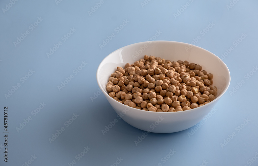 Selected grains of raw chickpeas in a white bowl on a blue background. Horizontal orientation, copy space, no people Selected grains of raw chickpeas in a white bowl on a blue background. Horizontal orientation, copy space, no people