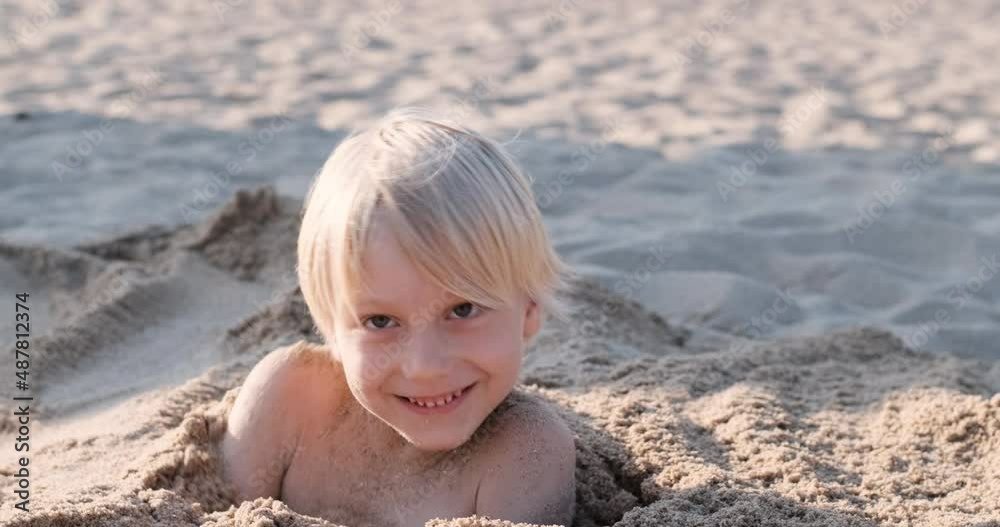 Smiling blond boy is fooling around on the beach. Child is buried in the sand, getting out of the sand pit. Holiday with children on the sea. Smiling blond boy is fooling around on the beach. Child is buried in the sand, getting out of the sand pit. Holiday with children on the sea.