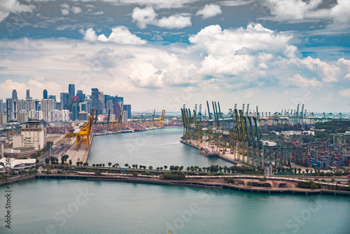 The huge busiest logistic port in Singapore, plenty of cranes to move containers, huge cargo ships in the background, skyscrapers on background