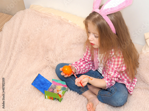A little blonde girl with bunny ears paints an Easter egg with a paintbrush. Easter preparation. Making Easter decor.