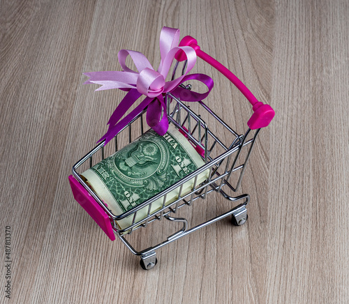 small market trolley with a coin dollar banknote on a wooden gray background close up, supermarket handcart with money photo