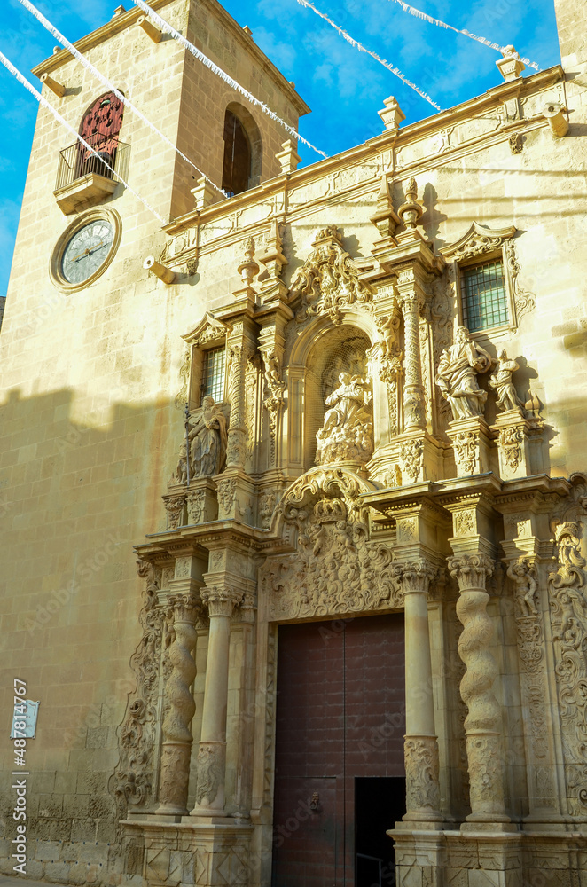 Artful entrance of the Basilica of St Mary of Alicante between the clock tower and another tower under clear blue sky. Valencian Gothic style from the 14th and 16th centuries.