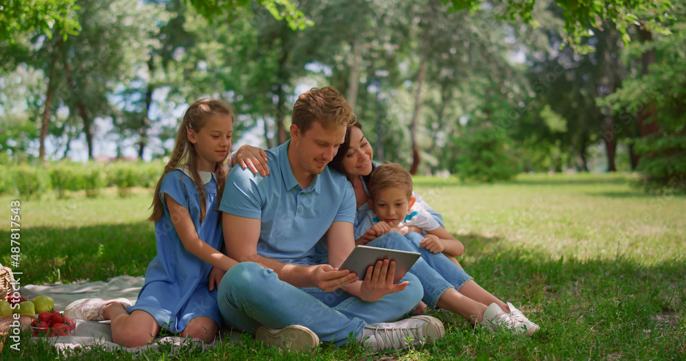 Young family sitting with tablet on sunny park. Happy people use laptop outdoors