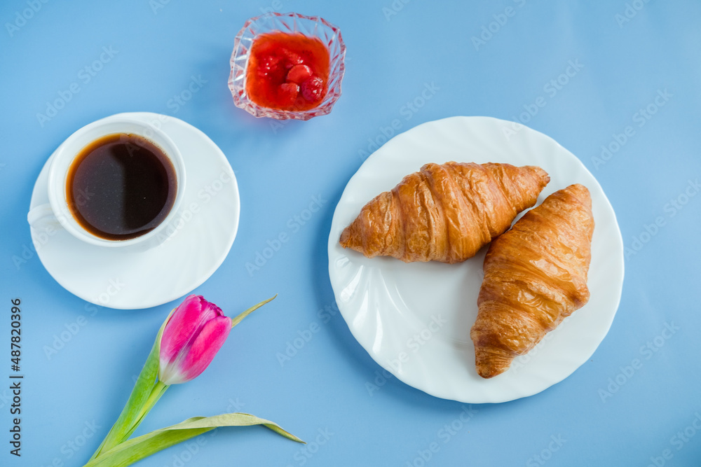 Croissants with jam, pink tulip and morning coffee in a white cup on a blue background.