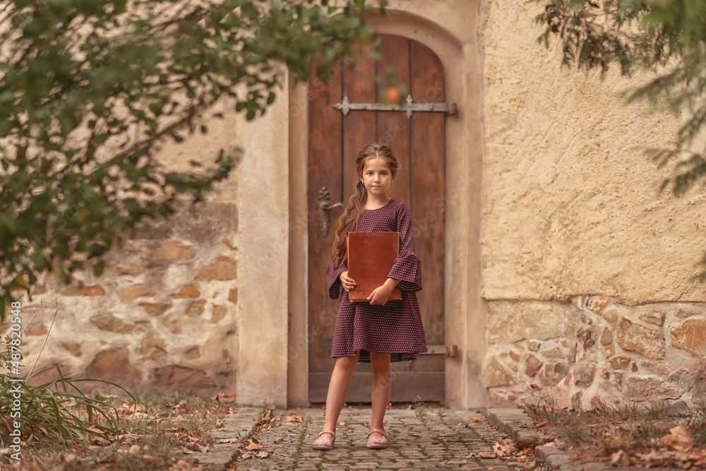 Portrait of a beautiful little girl with an old encyclopedia in her hands in the courtyard of the house.