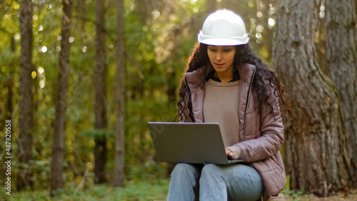 Millennial woman young experienced specialist forestry engineer environmentalist technician in hardhat checking trees entering data into laptop taking reforestation measures observing nature reserve photo