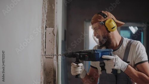 Builder drilling a wall with hammer drill, wearing safety glasses and headphones. Powerful hammer drill breaks brick. Sun breaking through window of construction studio High quality 4k footage photo