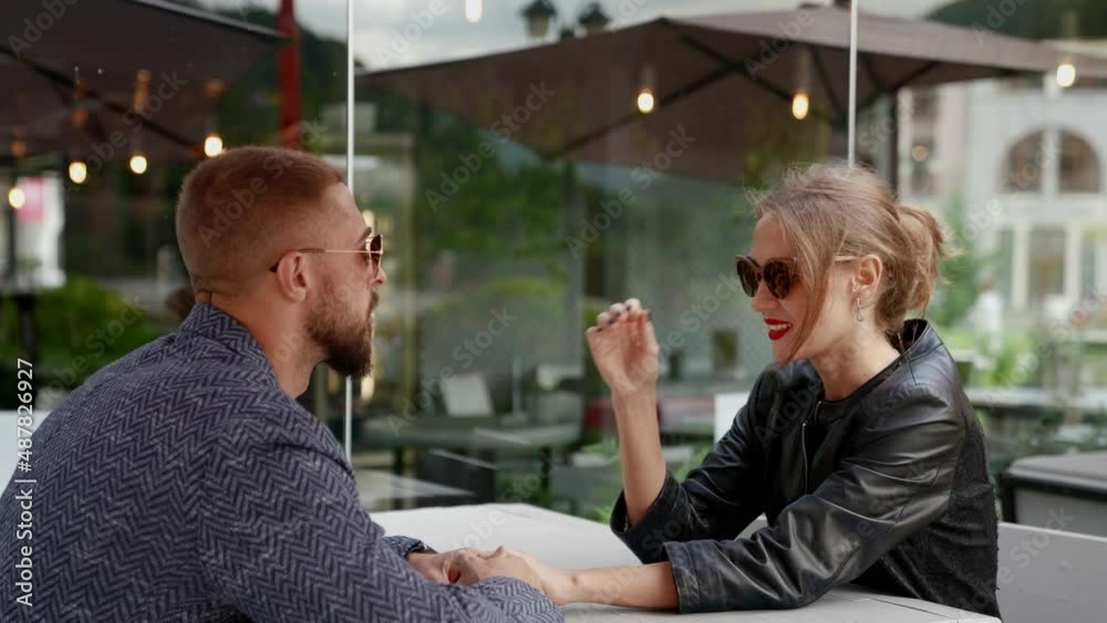 a couple is sitting at a cafe table on the street. a man with a beard and a blonde look at each other, then take off sunglasses. the camera is moving a couple is sitting at a cafe table on the street. a man with a beard and a blonde look at each other, then take off sunglasses. the camera is moving