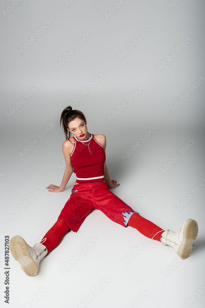 full length of young woman in red sportive outfit and boots sitting on grey.
