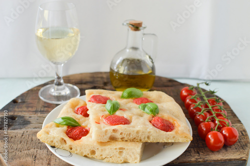 Italian sourdough focaccia bread with tomatoes and basil on the white plate and wooden board with cherry tomatoes and olive oil and white wine top view