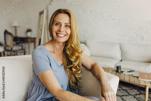 ttractive adult woman sitting on coach in living-room with loft design, having beautiful, clean and combed wavy hair, big blue eyes matching with color of her dress. Natural beauty concept