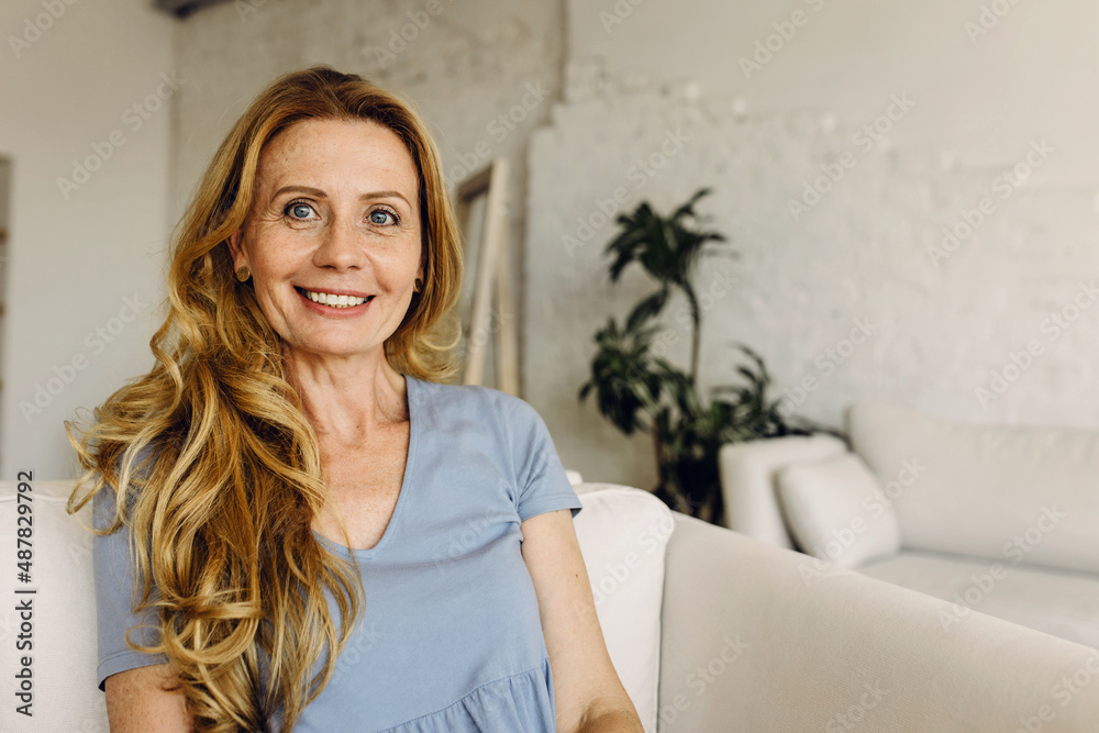 Smiling mature woman sitting on sofa against white home interior background, looking happy, smiling widely, looking at camera, having rest after house work, dressed casually in blue clothes Smiling mature woman sitting on sofa against white home interior background, looking happy, smiling widely, looking at camera, having rest after house work, dressed casually in blue clothes