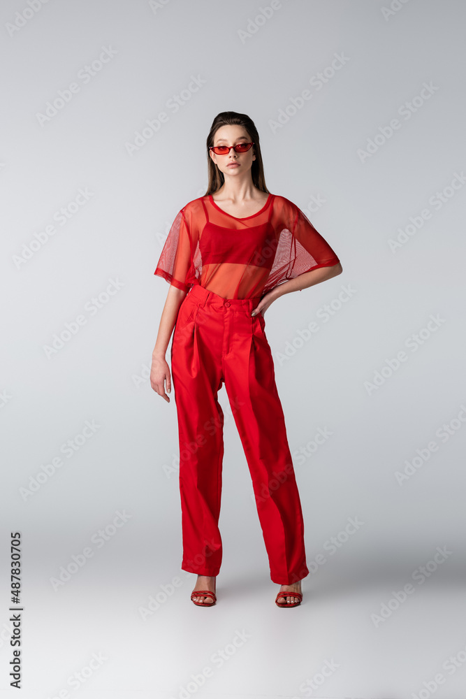 full length of young model in sunglasses and red trendy outfit posing with hand on hip on grey. full length of young model in sunglasses and red trendy outfit posing with hand on hip on grey.