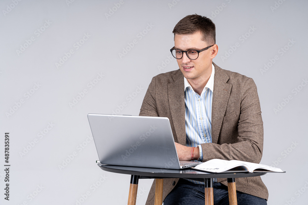 Handsome businessman sitting at the table and working at the laptop. Young male in suit is sitting and looking at the screen. Full length portrait Handsome businessman sitting at the table and working at the laptop. Young male in suit is sitting and looking at the screen. Full length portrait