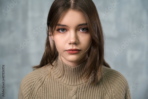 Studio portrait of young beautiful girl with doll face. Grey wall in background. Knitted sweater.