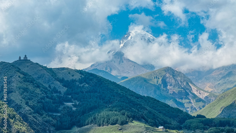 Distant view on Gergeti Trinity Church in Stepansminda, Georgia. The church is located the Greater Caucasian Mountain Range.Clouds are covering the snow-capped Mount Kazbegi in the back. Hiking trail Distant view on Gergeti Trinity Church in Stepansminda, Georgia. The church is located the Greater Caucasian Mountain Range.Clouds are covering the snow-capped Mount Kazbegi in the back. Hiking trail