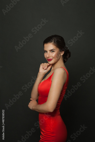 Stylish woman with makeup and trendy hairdo in red silky dress smiling on black background.