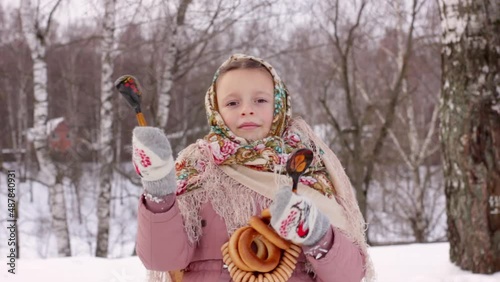 Cute girl in a traditional Russian headscarf and mittens playing on spoons for Maslenitsa festival on winter background. Closeup portrait of a child for Srovetide holiday. photo