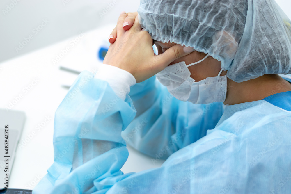 Fototapeta premium A tired female doctor wearing face mask and surgical overalls is sitting at her desk in the medical office with her eyes closed.Healthcare and medical concept.