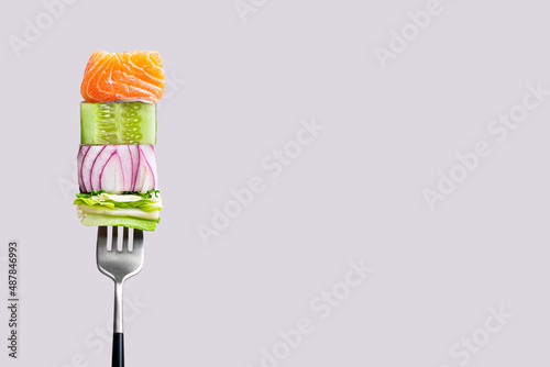 Close-up of fork with food on it: delicious fillet salmon, cucumber, onion, green salad on gray color background. Concept of healthy diet and clean eating with fish and vegetables, balanced nutrition