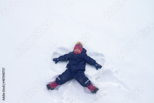 A Caucasian baby girl in a blue winter jumpsuit and a knitted hat lies on the snow and portrays an angel in winter